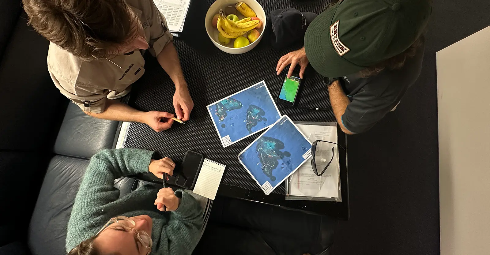 A bird's-eye view photo of three scientists sitting around a small table in a boat cabin. They are studying satellite photos of a series of small islands.