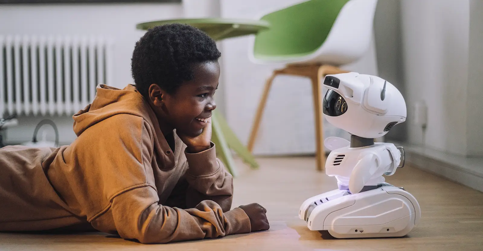 A boy of young middleschool age lies on the floor on his stomach, with his head resting on one hand. He is smiling happily and looking at a small white robot. The robot is small, with an oval head, a small body with paddles for arms, and a set of treads for locomotion.