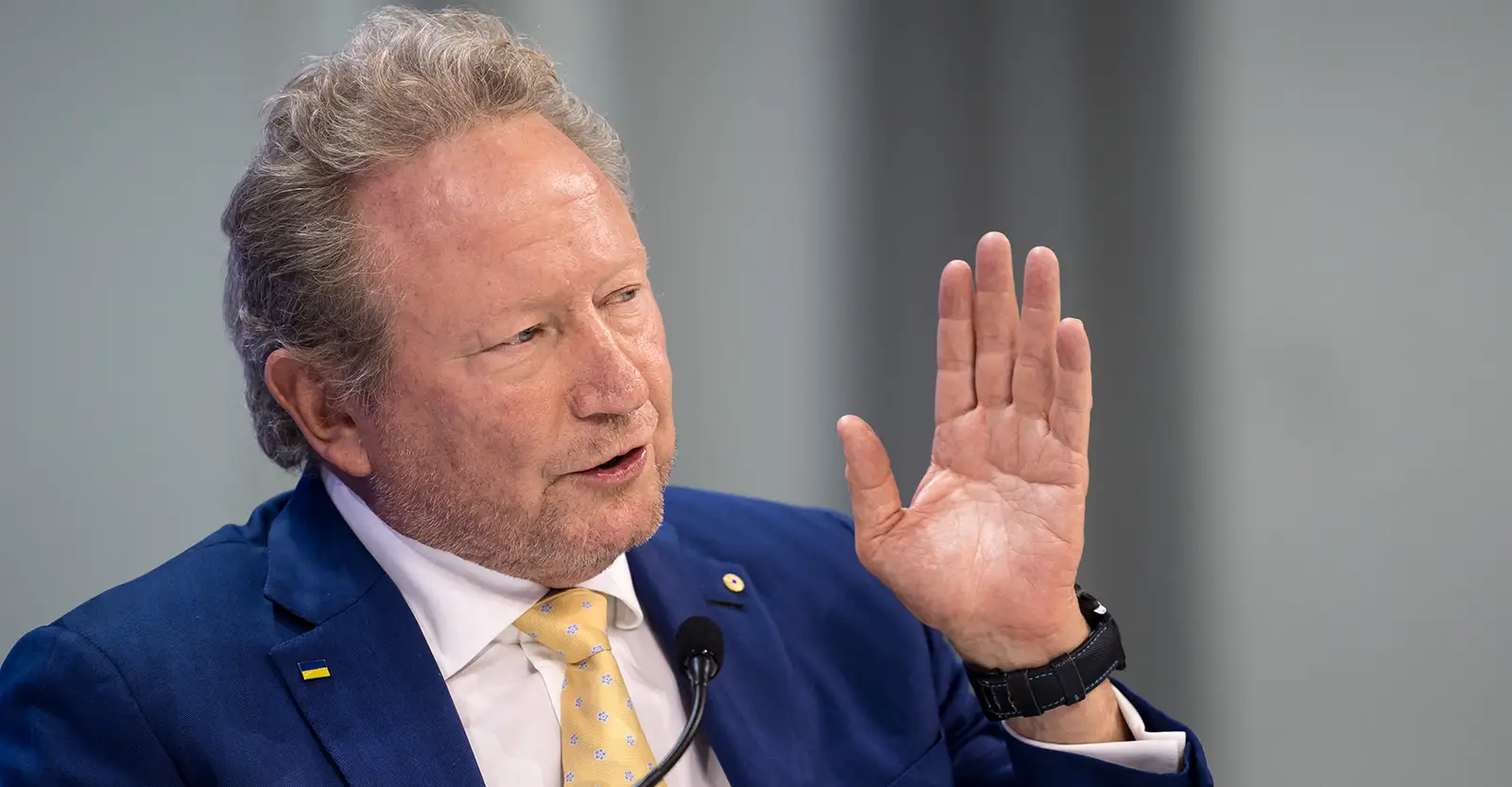 Andrew Forrest gestures with his hand held up while speaking into a podium microphone. He is wearing a blue suit jacket, white shirt and yellow and light blue patterned tie. He wears a pin of the Ukraine flag on one of his lapels.