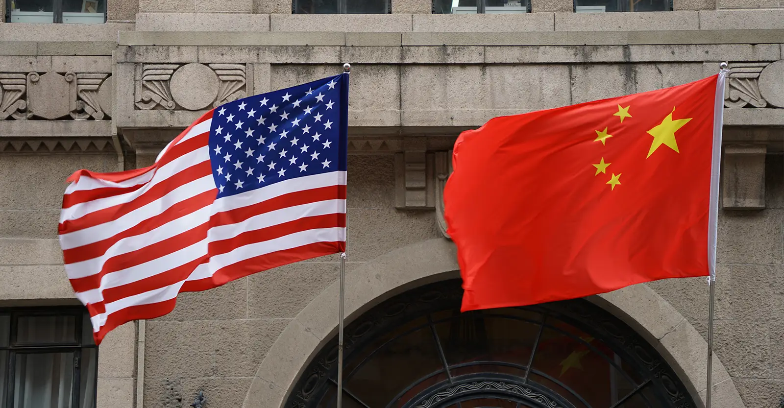 The US flag and the Chinese flag, flying towards the left, in front of a grey stone wall of a multi-storey building.