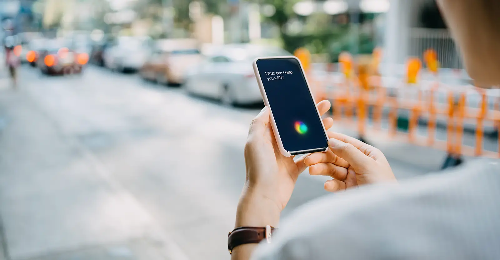 A midshot of a woman standing next to an urban street, holding a phone with the words 'What can I help you with?' on it. The photo is taken over the woman's shoulder and has a shallow depth of field so only the hands and phone are in focus.