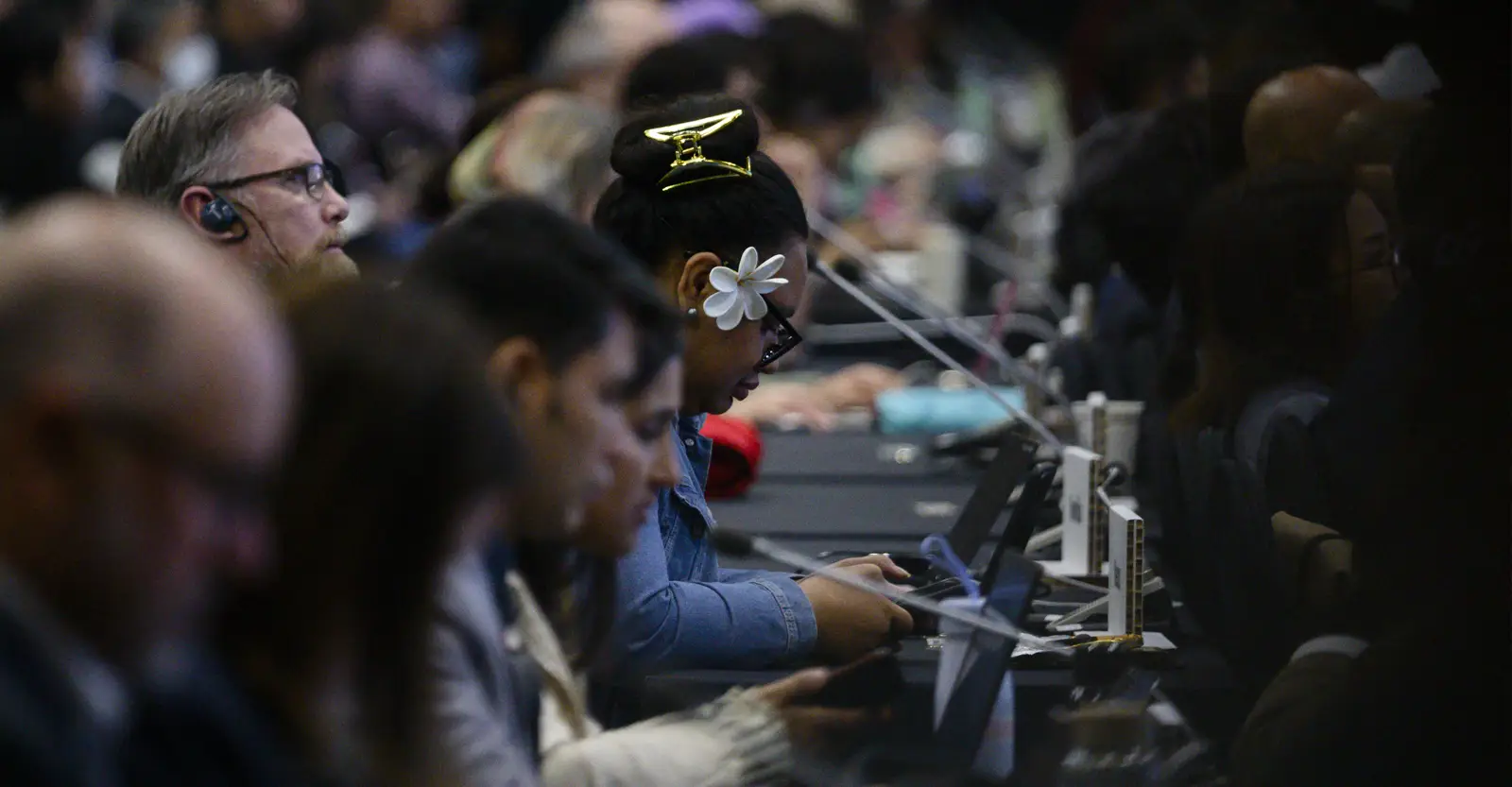 A side view of a diverse group of people sitting at tables that have microphones on them. Some are looking at devices translating for them, others have earpieces for live translation.