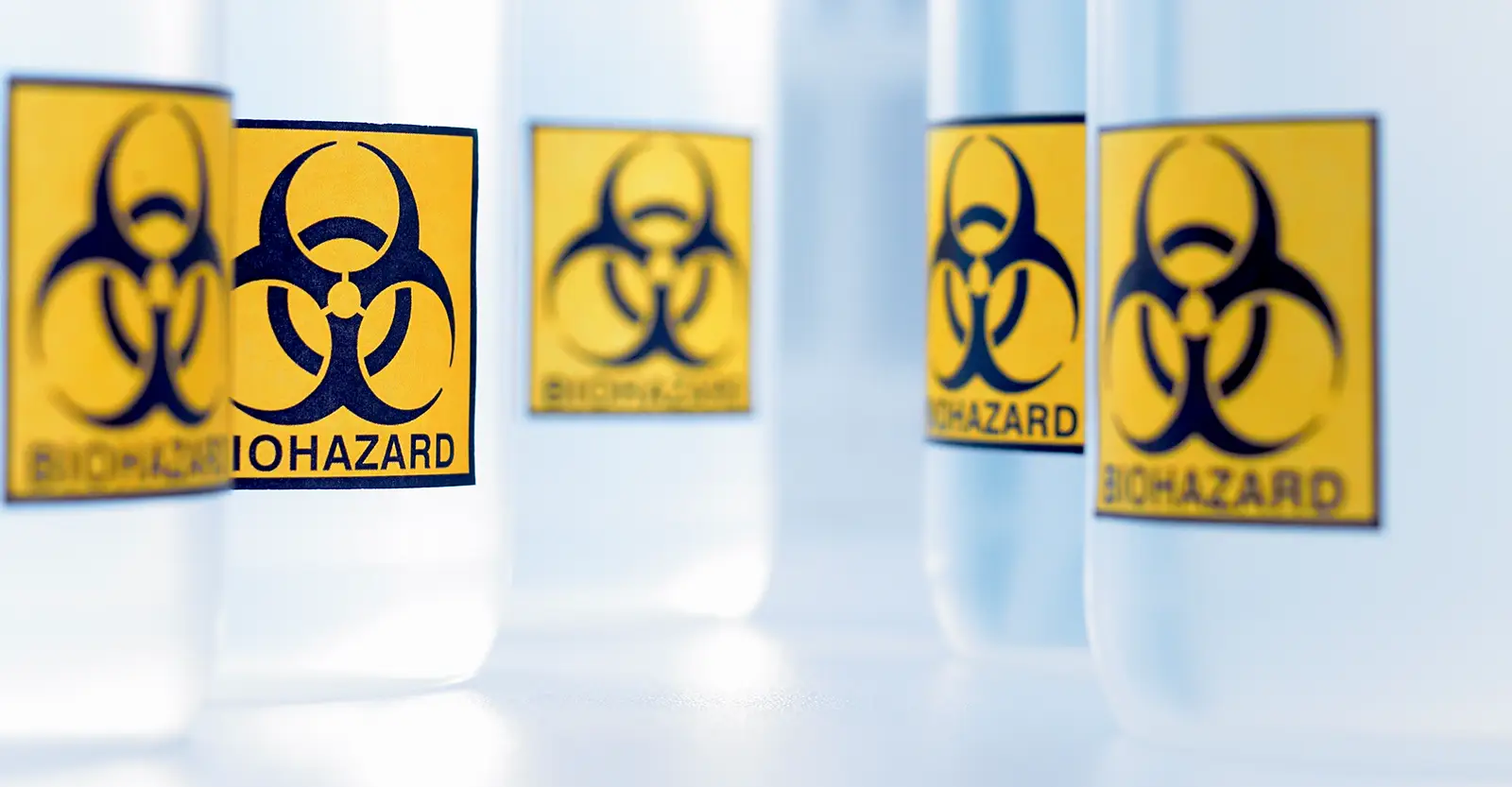 Wide-mouthed plastic bottles filled with clear liquid and with yellow biohazard warning labels on them are lined up in a lab.