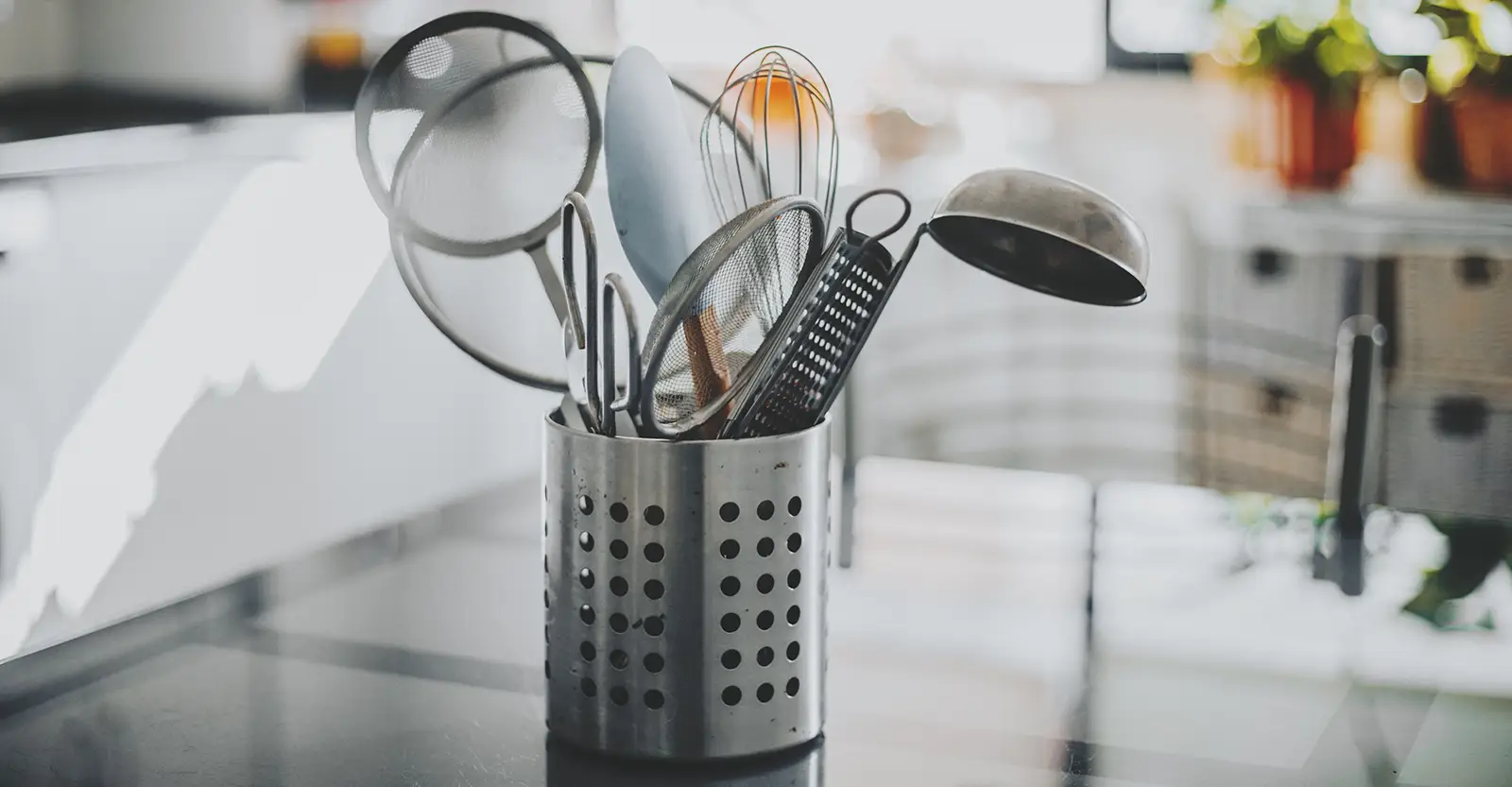 A metal utensil holder sites on a glass table, filled with a variety of stainless steel cooking utensils.