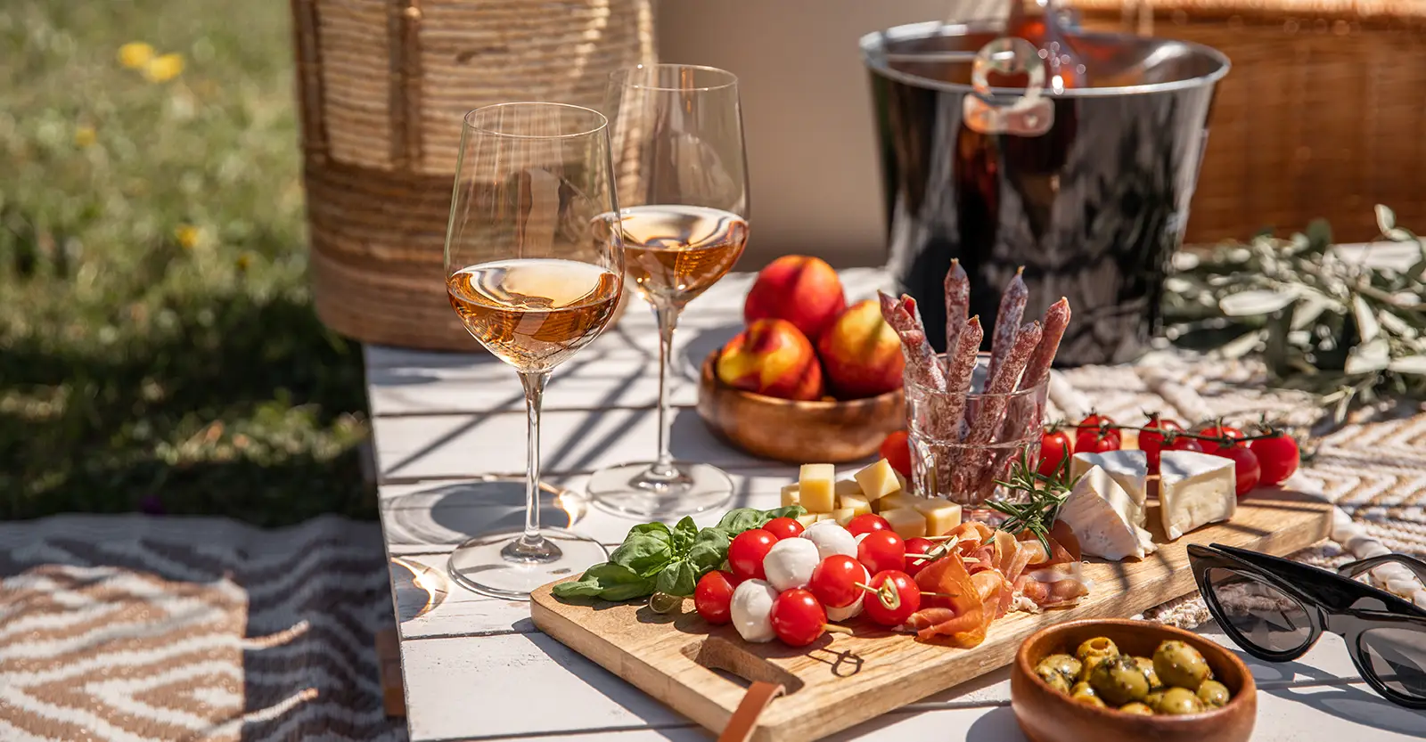 A picnic table in a sunny field, with a cheese platter, some antipasto, fruit, two glasses of rosé and a wine cooler with a full bottle of rosé in the background.