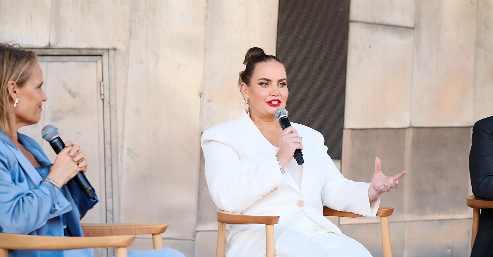 Jelena Dokic, wearing a white suit, speaks into a hand-held microphone. She is sitting, and making an gesture for emphasis with her other hand. Two her right is Shaynna Blaze, wearing a blue suit and watching Jelena with a smile. To her left, only the arm of another panel member can be seen.