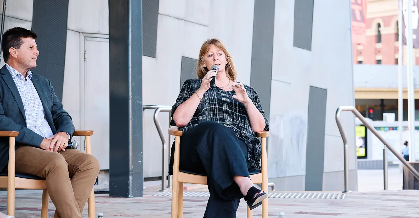 Janise Mitchell sits on a chair, speaking into a hand-held microphone. She is wearing black slacks and a black and grey wide plaid pattern top. To her right, also sitting in a chair is panel member Paul Oliver, wearing tan trousers, a light blue shirt and a drak blue jacket. They are outside and in the background can be seen a central urban street with traffic lights.