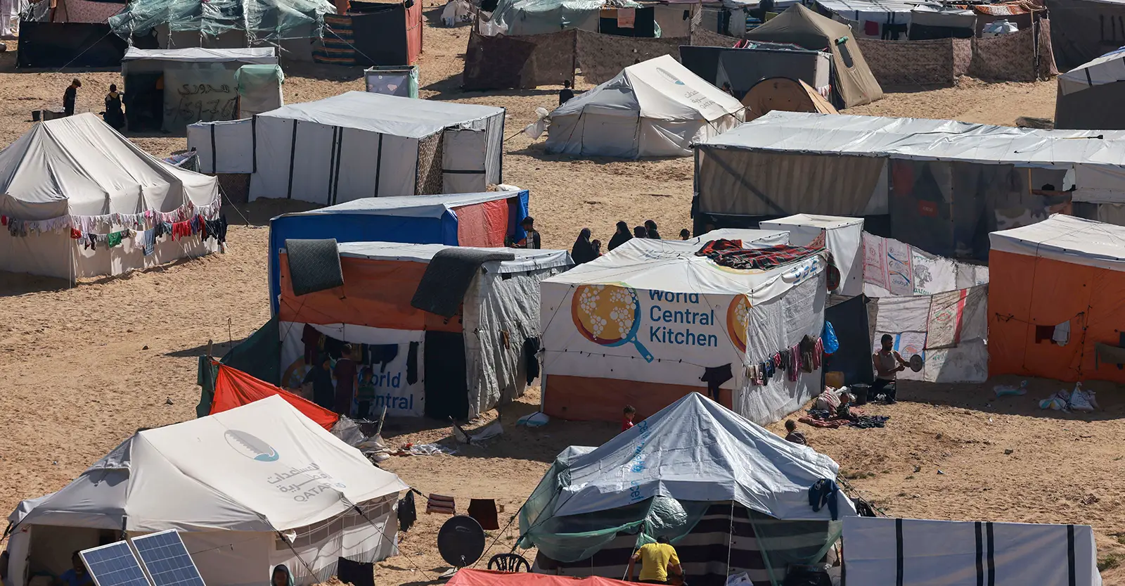 A long shot of a temporary camp, built on bare earth and made up of various kinds of tents. In the middle of the shot there is a tent with the logo of World Central Kitchen on it.