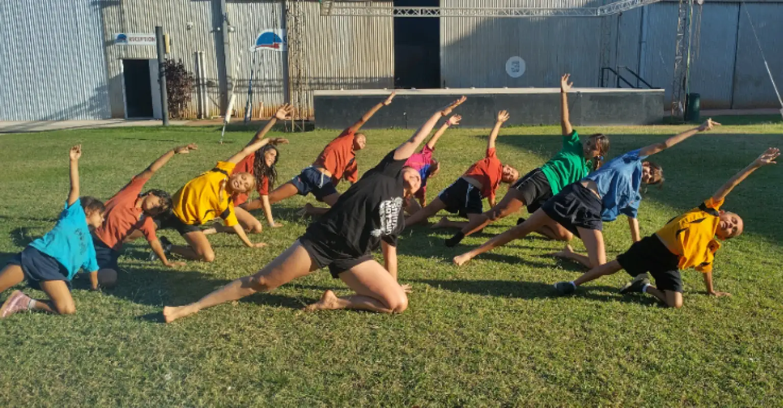 Tara Gower teaching dance to a group of Indigenous children wearing brightly coloured t-shirts. They are all kneeling on one knee and stretching their opposite hands over their heads.