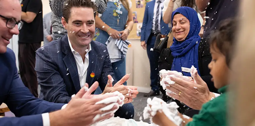 Four adults, including John Hartman, playing with a small girl at an early learning centre. They are all clapping their hands together to make shaving foam fly, and it looks like a happy, noisy play time.