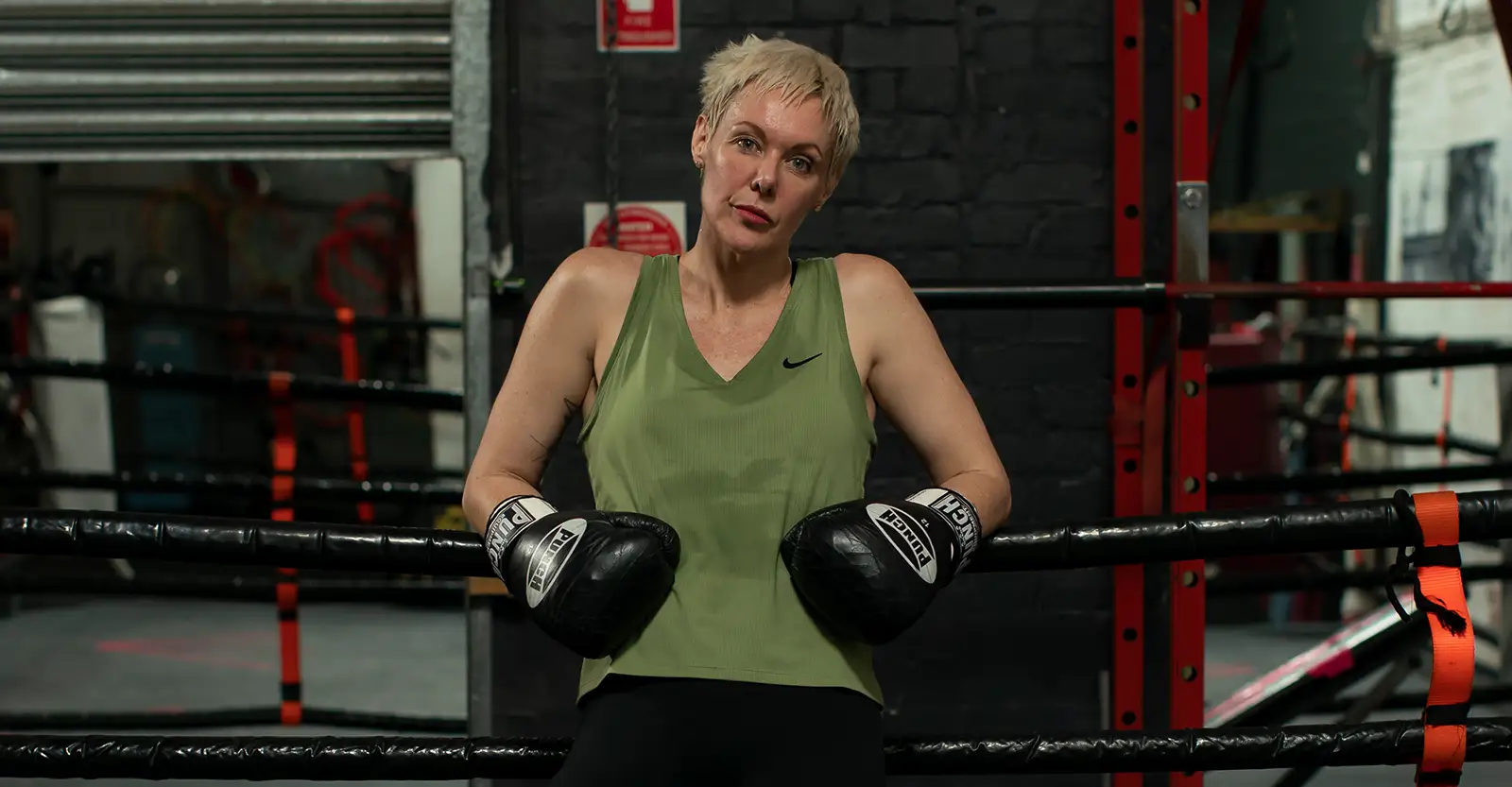 Donna Lyon leans against the ropes of a boxing ring in a gym, wearing work-out clothes and black boxing gloves. She looks powerful and determined. 