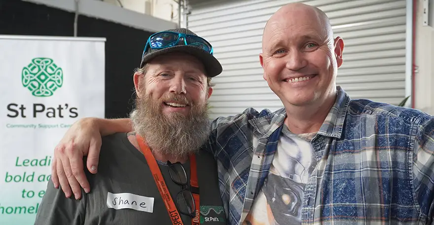 A mid-shot of Jaron smiling at the camera, with his right arm around Shane’s shoulders. Jaron is tall, with a shaved head and blue eyes. Shane is a bit shorter than Jaron and sports a long, bushy light-brown and grey beard. He is also smiling at the camera.