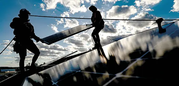Darkly shadowed silhouettes of two workmen about to install a solar panel on a roof. In the foreground leading to the men are the previously installed panels and in the background is a brightly lit blue but partly clouded sky.
