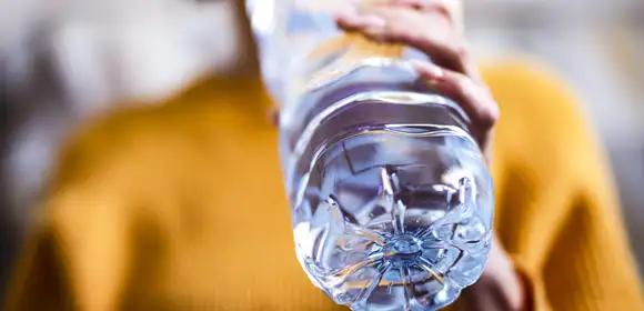 A woman wearing a bright mustard jumper holds a large plastic water bottle up to her mouth. The bottom of the plastic bottle is in focus and the image has a shallow depth of field so the rest of the photo is in various stages of blur.