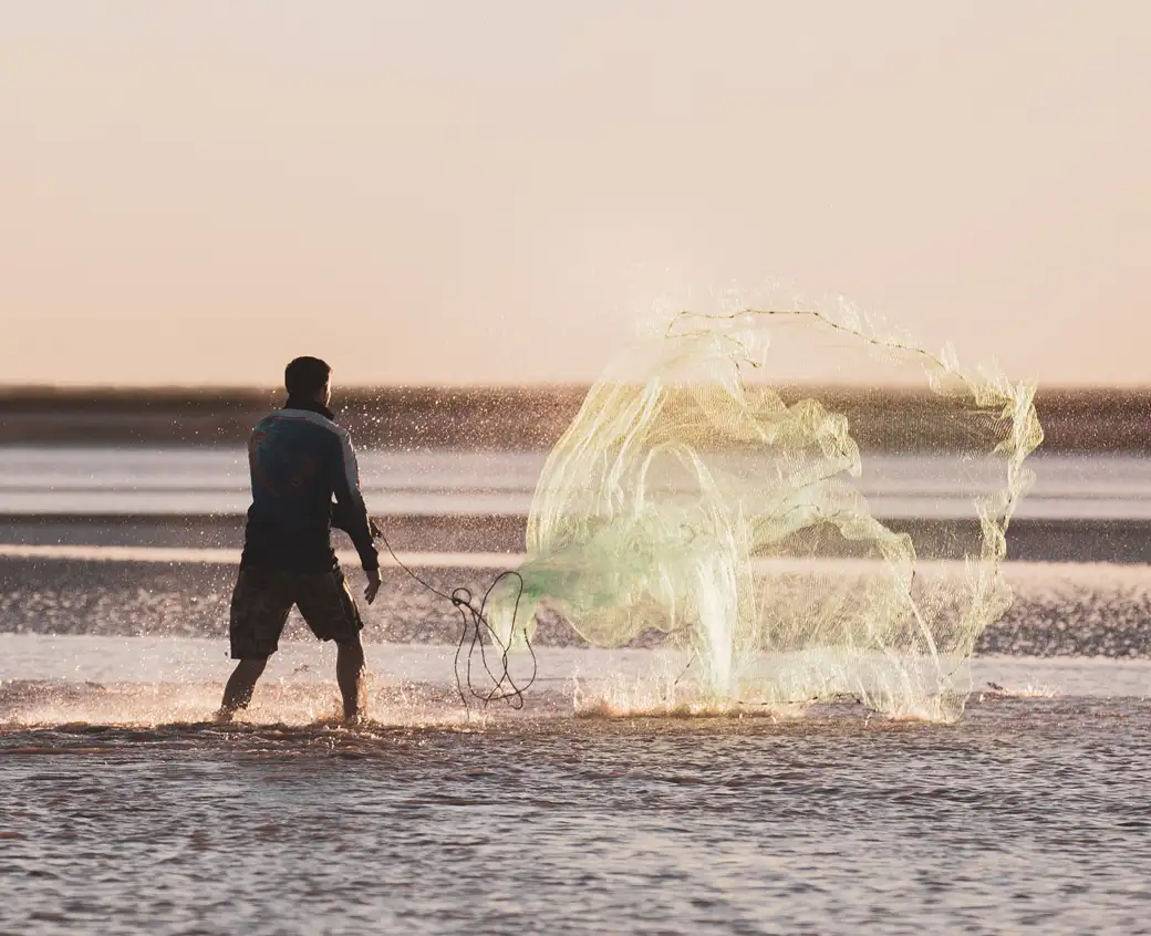 A man standing in ankle deep water casts a round fishing net into the sea. The photo has captured the moment as the gathered net begins to spread out mid-air. The man is facing away from the camera, towards the rising sun, which is giving the photo an orange-pink tint.