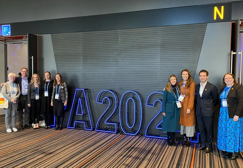 A long shot of Minderoo Foundation staff at the 2024 Philanthropy Australia conference posing in front of tall blue neon light up letters that say 'PA2024'. The characters P and 4 are obscured by people standing in front of them - to the left, four women and three men; to the right, three women and one man. They are all wearing business casual attire and conference lanyards.
