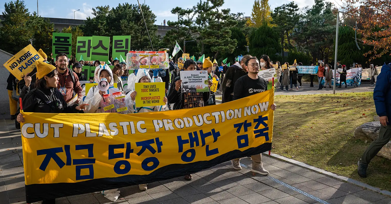 A group of protesters march through a pedestrian area carrying banners and signs in both Korean and English. At the front of the procession a bright yellow banner reads ‘Cut plastic production now’. In the background are a number of trees.