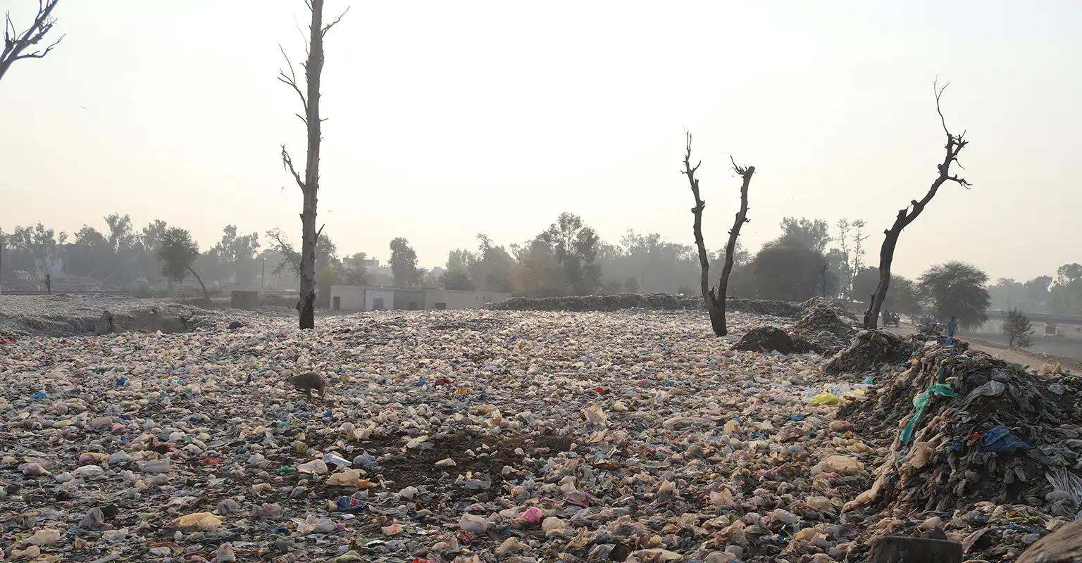 A large field is covered in plastic rubbish, spread over the entire surface, and heaped up in mounds. In the background can be seen square single storey buildings, and trees. The sky is hazy and a light orange colour.