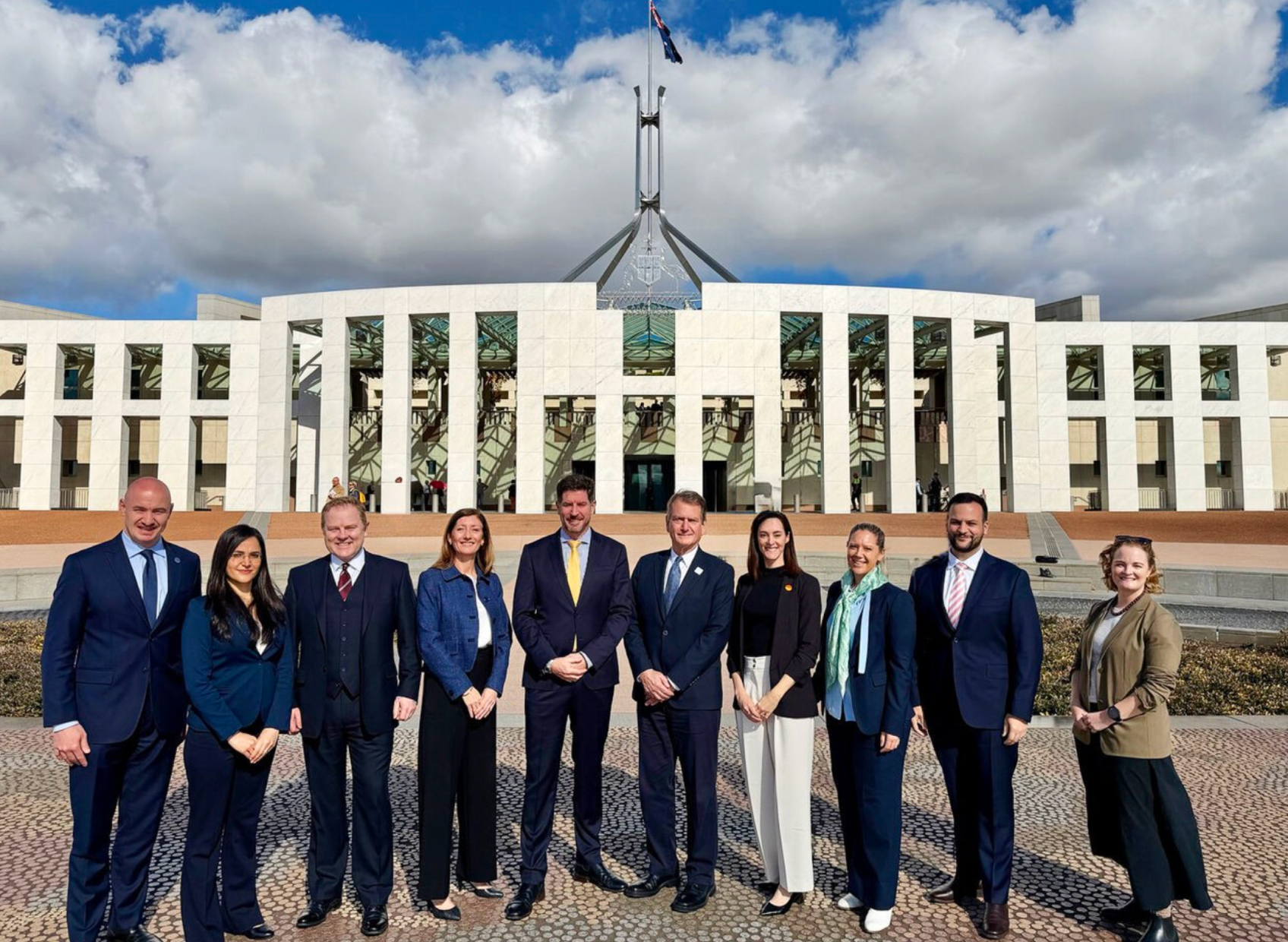 A group of men and women in professional wear stand outside Australia's Parliament House in Canberra, smiling into the camera.