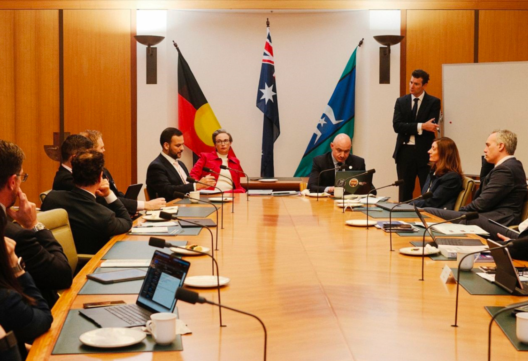 A large group of men and women in business attire are seated around an executive boardroom table with microphones at each seat. In the background against the wall are the Australian Aboriginal flag, the Australian national flag and the Torres Strait Islands flag