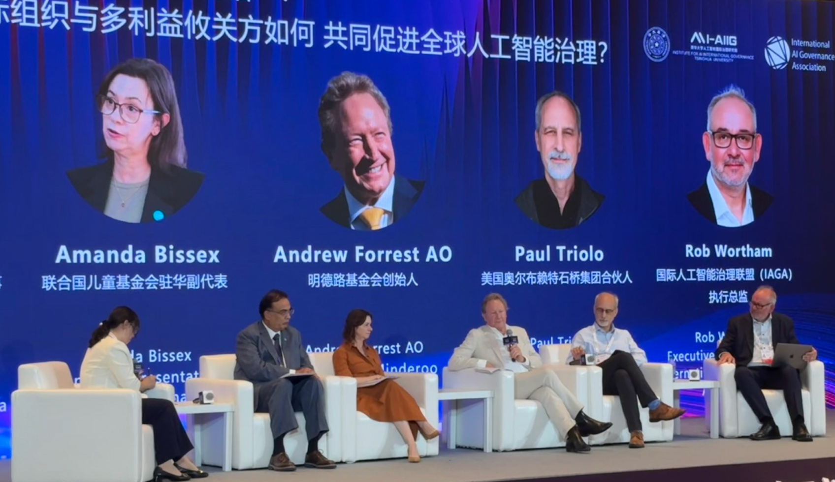 A group of two women and four men sit in comfortable chairs on a stage with a huge screen behind them. The screen is a slide with the speaker's details on them in both English and Chinese.