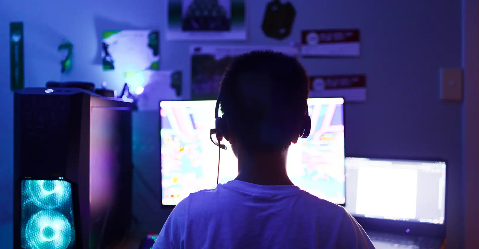 This image is shot from directly behind a boy sitting at a computer with a headset on. Set up on the desk in front of him is a computer tower with light-up fans, a desktop monitor, and a laptop.