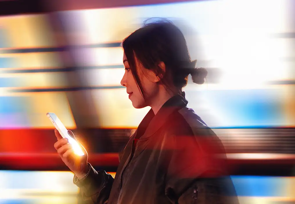 A side on view of a young woman with dark hair, looking at a phone in her right hand. The background is stripes of bright lights that have been blurred, and there is motion blur around the young woman.