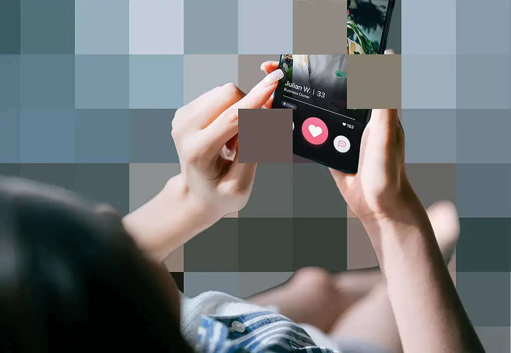 A birds eye view of a woman holding a mobile phone. On the screen of the phone is a profile on a dating app. There are small coloured squares over much of the screen, giving a pixellation effect.