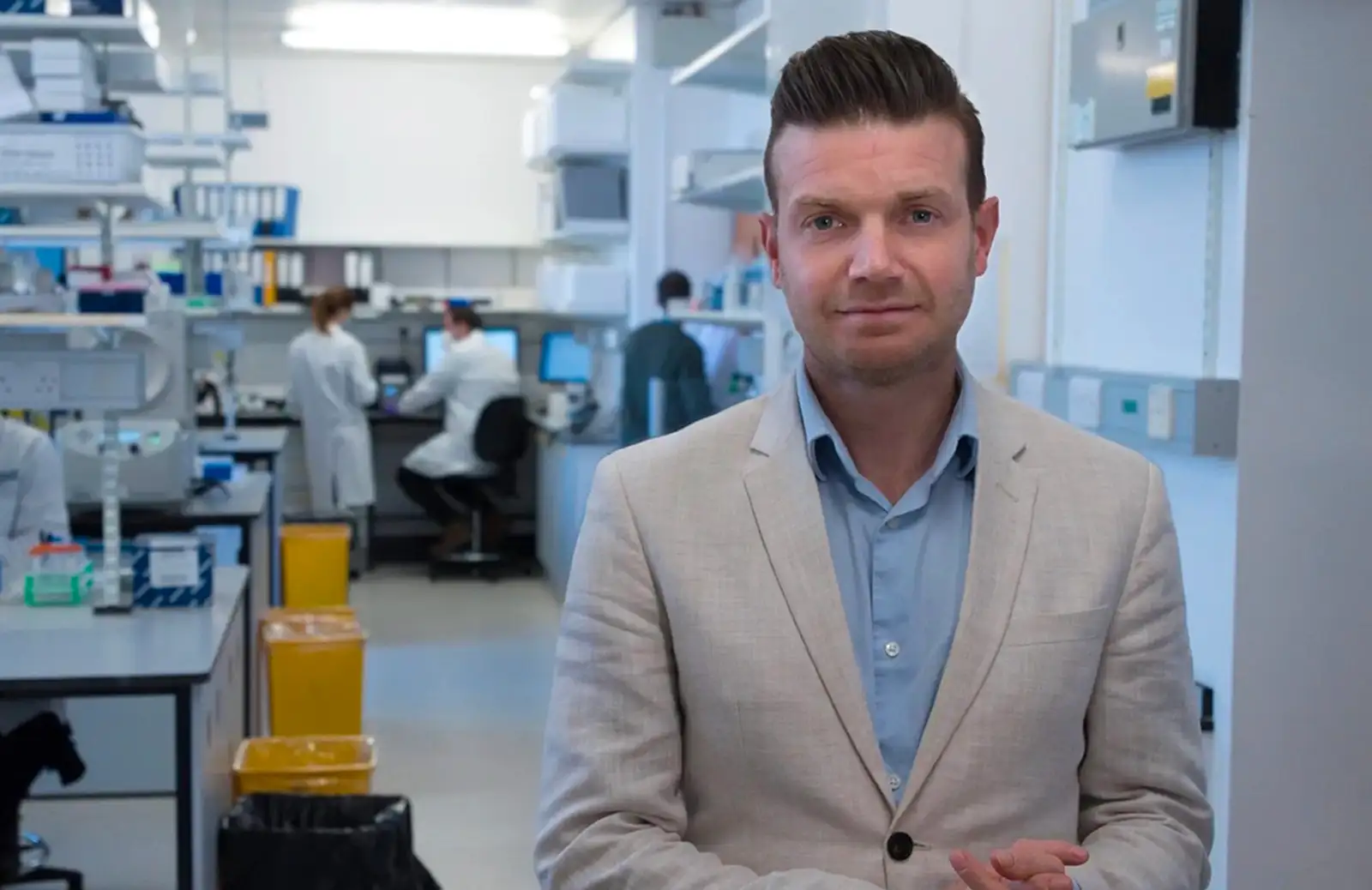 Professor Richard Mair stands in a lab, wearing a blue shirt and a beige linen suit coat. In the background can be seen some researchers, most of whom are wearing lab coats.
