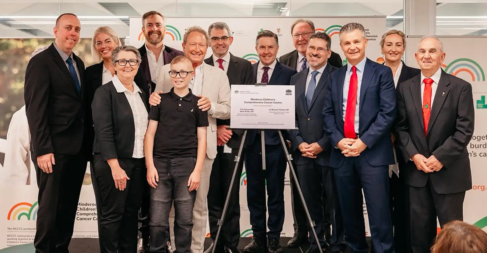 A group of people, mostly middle aged men in suits, are grouped around a plaque announcing the opening of the Minderoo Children’s Comprehensive Cancer Centre. There is a young teen boy at the front of the picture.