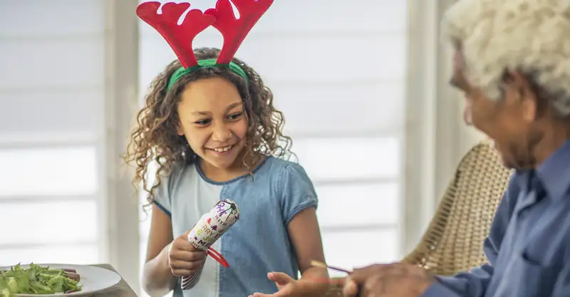 A happy young First Nations girl wearing red reindeer antlers on a green headband has just won a Christmas cracker after having pulled it with her grandfather.