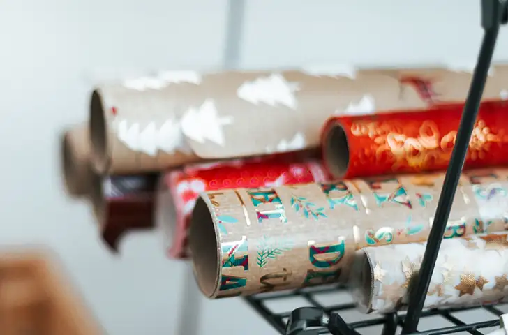 A black wire shelf holding a number of rolls of Christmas wrapping paper, with metallic effect words or illustrations on them.