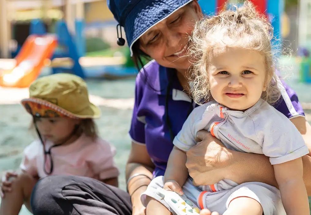 A group of primary aged children sit variously on grass and a blue esky, eating slices of watermelon. In the background, glimpses of their mums can be seen.