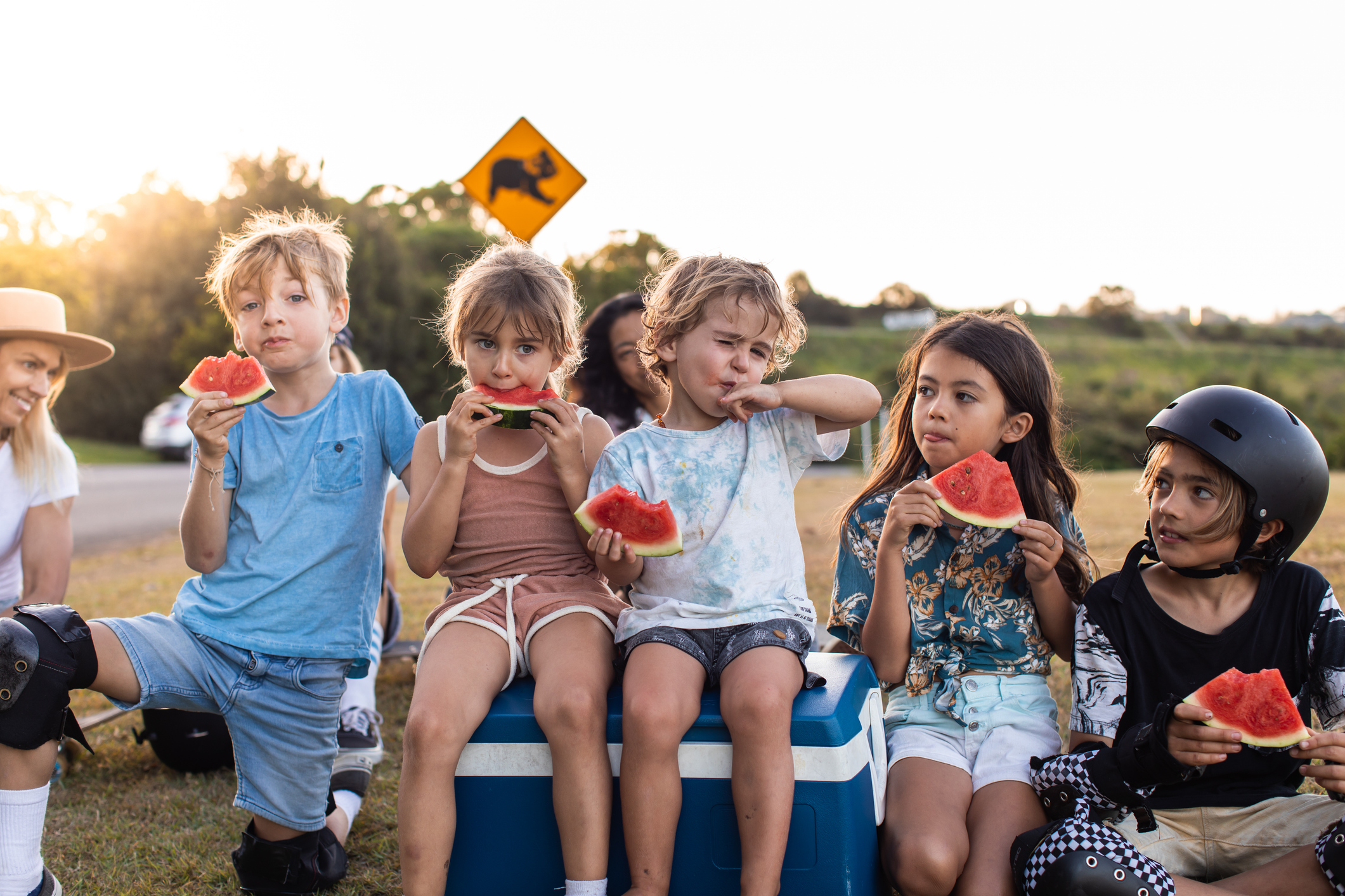 A group of primary aged children sit variously on grass and a blue esky, eating slices of watermelon. In the background, glimpses of their mums can be seen.