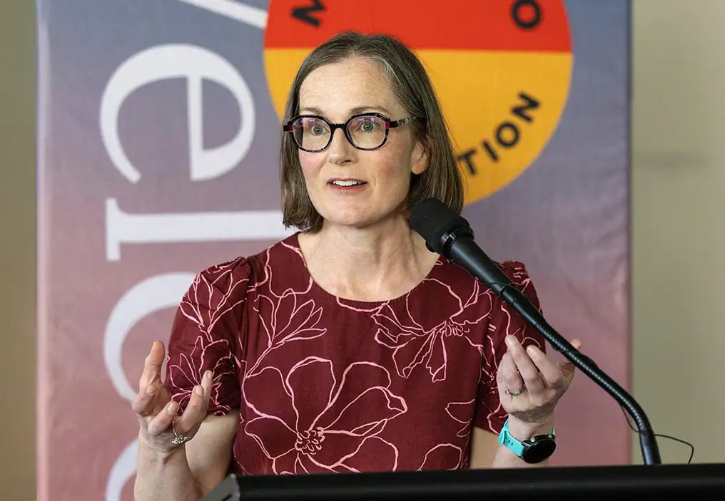 Caroline Croser-Barlow stands in front of a podium, with a pull up banner behind her. She is raising her hands in a gesture supporting her point. She wears a maroon top with a pattern of flowers outlined in a lighter colour and has solid rimmed glasses.