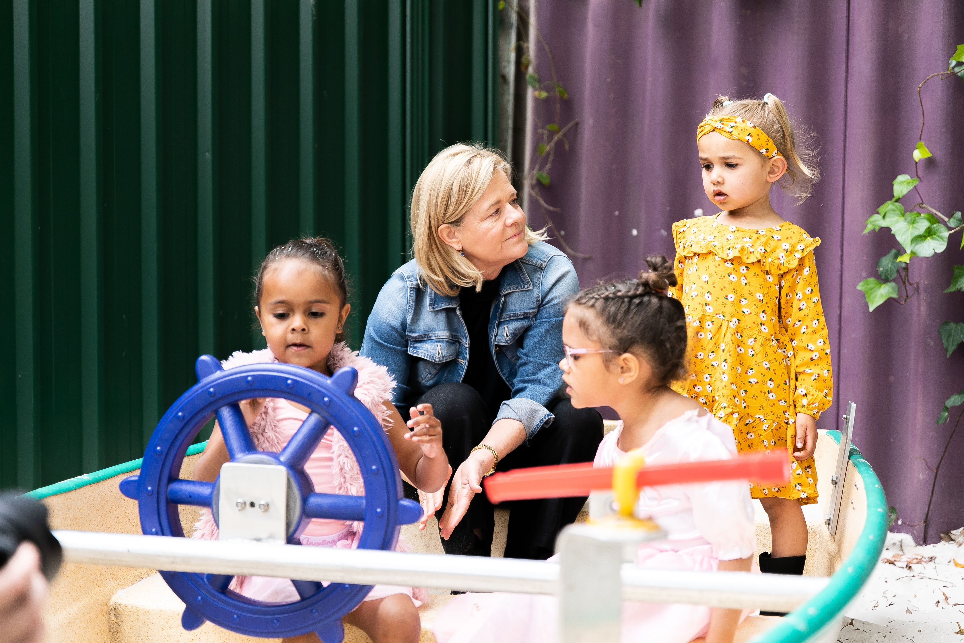 Nicola Forrest, wearing a denim jacket, sits in a play boat at a day care centre, accompanied by three young First Nations girls. One sits at the bright blue “wheel” of the boat while another watches her. The third is standing next to where Nicola sits, and is wearing a yellow ochre coloured dress with matching headband.