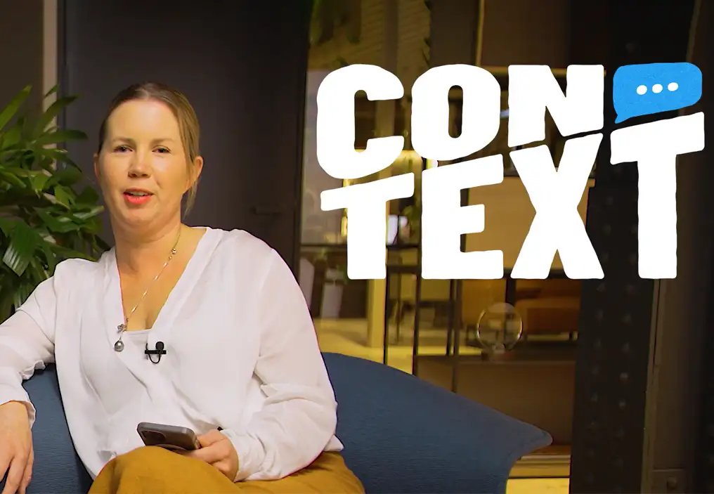 Amy Graham sits in a blue armchair, wearing a whit blouse and yellow ochre coloured slacks. She is holding a mobile phone and has a lapel mic fixed to her top. At top right, there is an overlay of the ConText logo.