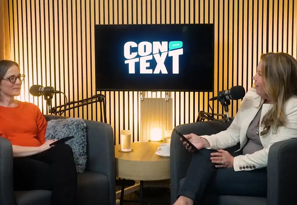 Two women sit in arm chairs in a podcast studio with slats of light coloured wood on the back wall. A television between them has the ConTEXT logo on it and they each have a large professional podcast mic on a long arm in front of them.