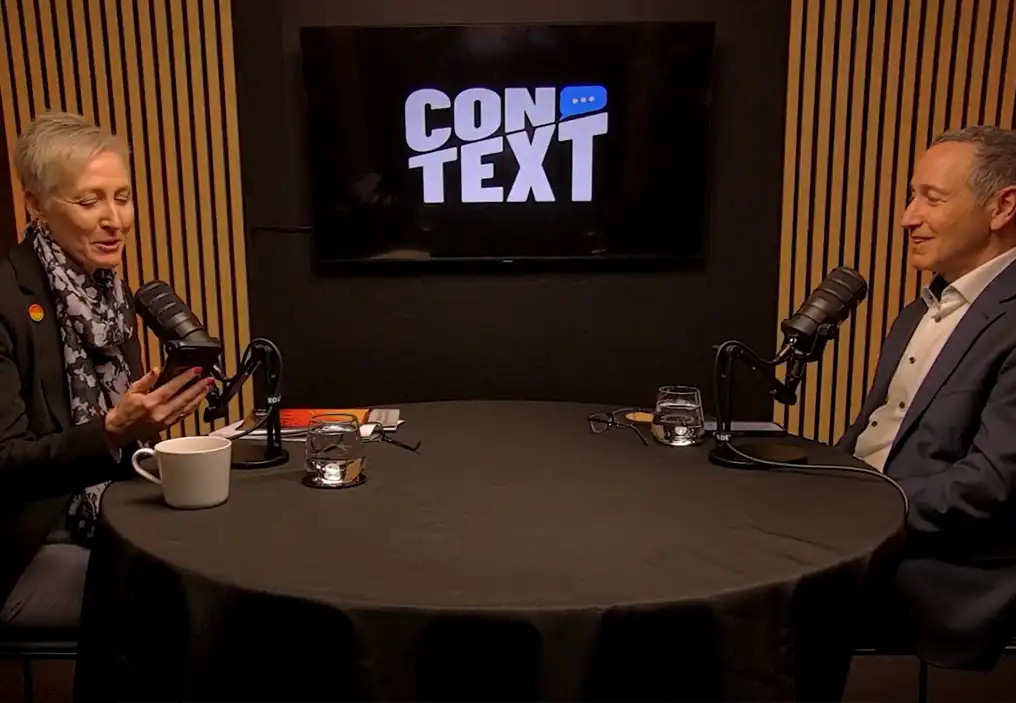 Dr Krista Singleton-Cambage and Dr Robert Glasser sit around a small round table in a podcast studio. On the wall behind, between them, there is a TV screen that has the ConTEXT logo on it.