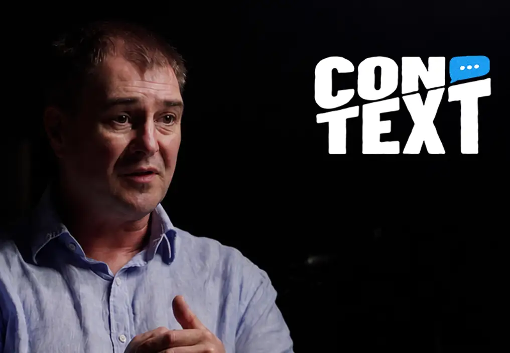 A still of Dr Kevin Thomas, wearing a blue business shirt and looking serious while gesturing with his hands. In the top right corner sits the ConTEXT logo.