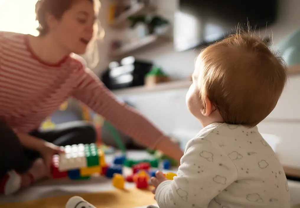 A young baby sits with their back to the camera, looking intently at their mother who is blurred in the background, reaching across to get some blocks to play with the baby.