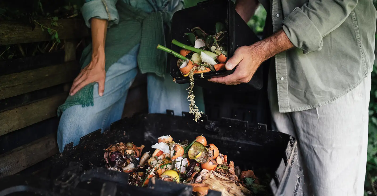 A shot of two people from the waist down to about knee height, standing next to a black plastic composting bin full of refuse, and emptying another black plastic container full of food scraps into it.