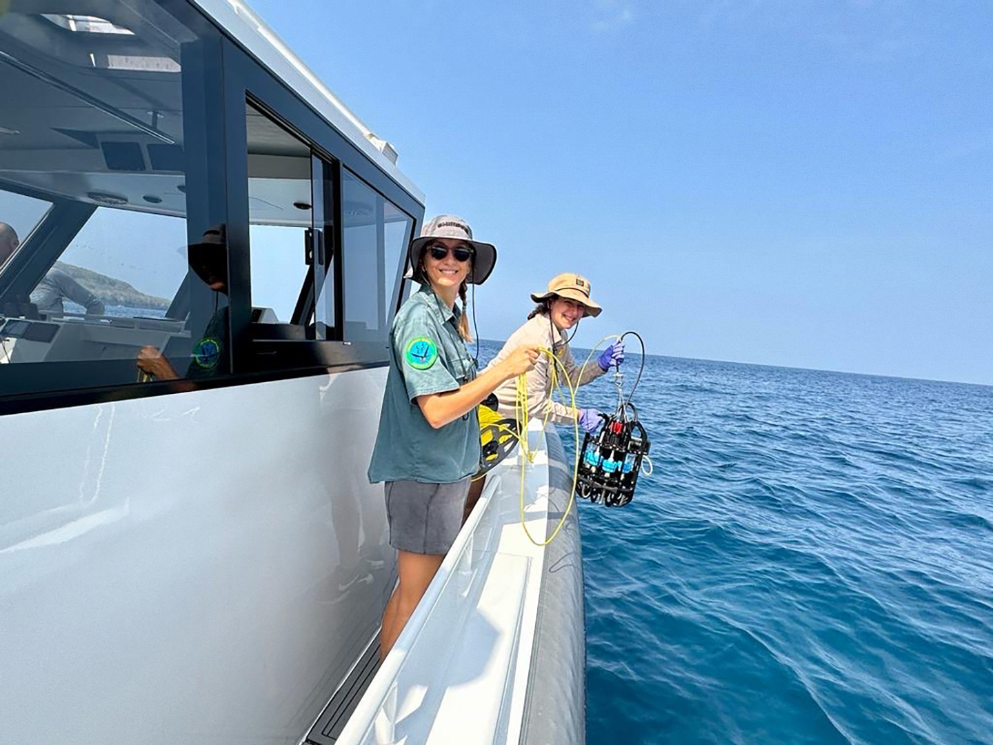 Two female scientist are at the side of a boat at sea, holding eDNA sampling equipment, ready to deploy it over the side. They are both smiling and looking at the camera.