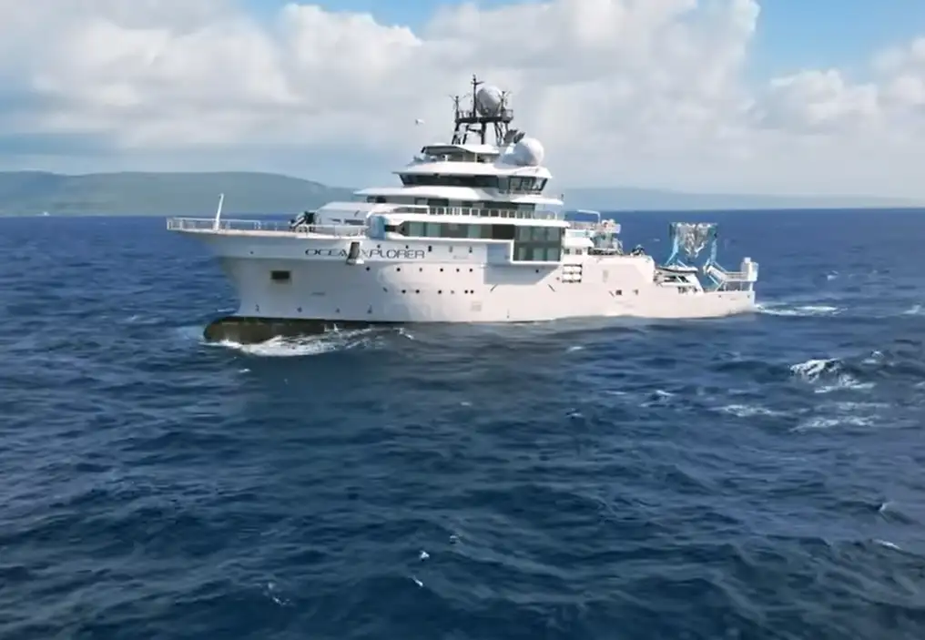 A large white and grey boat at sea, on a relatively calm deep blue ocean, but with land showing in the background.