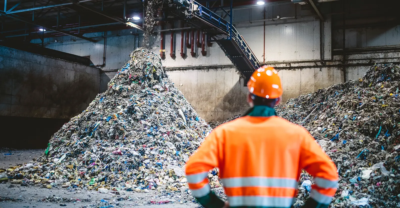 A worker wearing a bright orange safety jacket and an orange hard hat stands with his back to the camera, looking at waste falling from a conveyor belt in a rubbish processing factory.