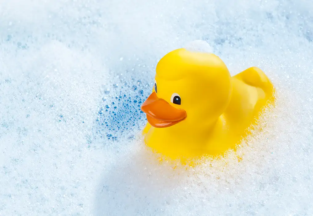A midshot of a yellow plastic duck with an orange beak floating on top of a bath full of white suds. The water below the suds is very blue.