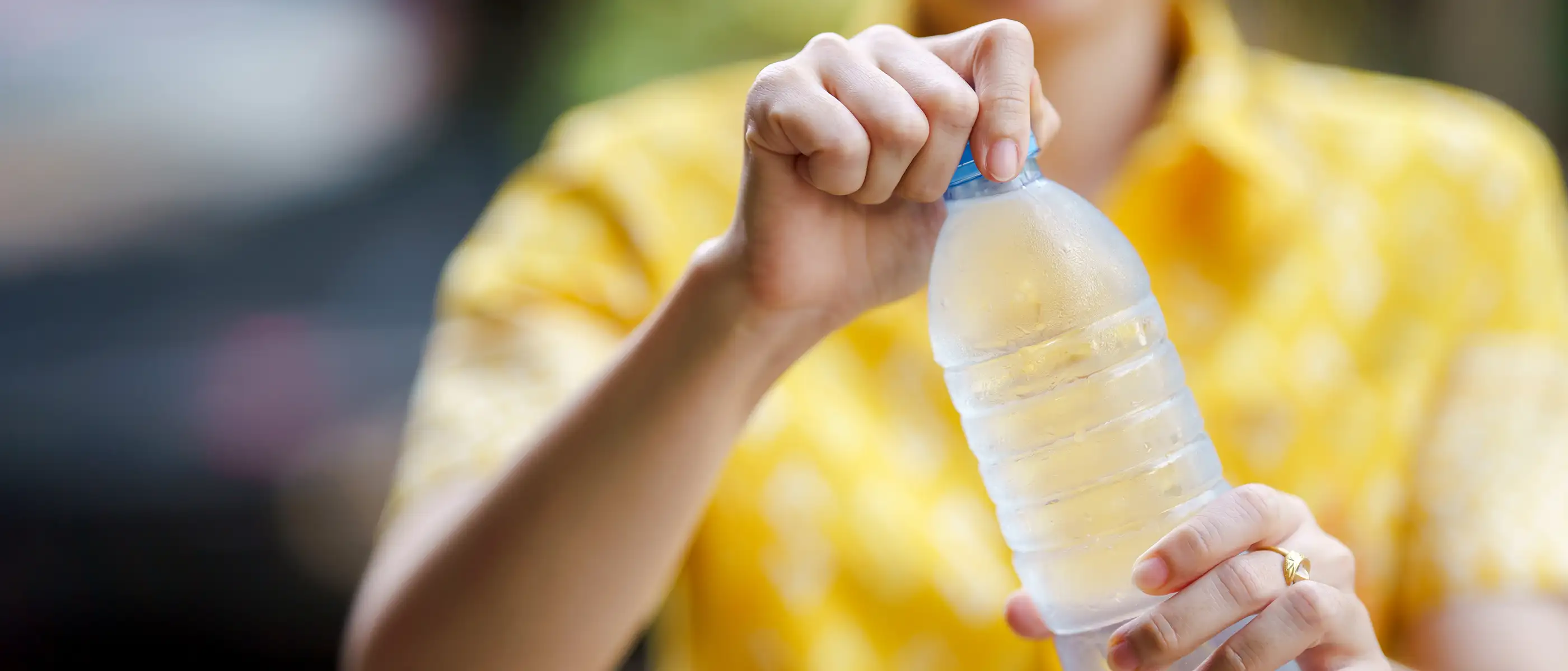 A close up of a single use plastic water bottle, covered in condensation. A woman holds the side and the cap of the bottle as though she is about to open it. She is wearing a yellow shirt and we cannot see her face.
