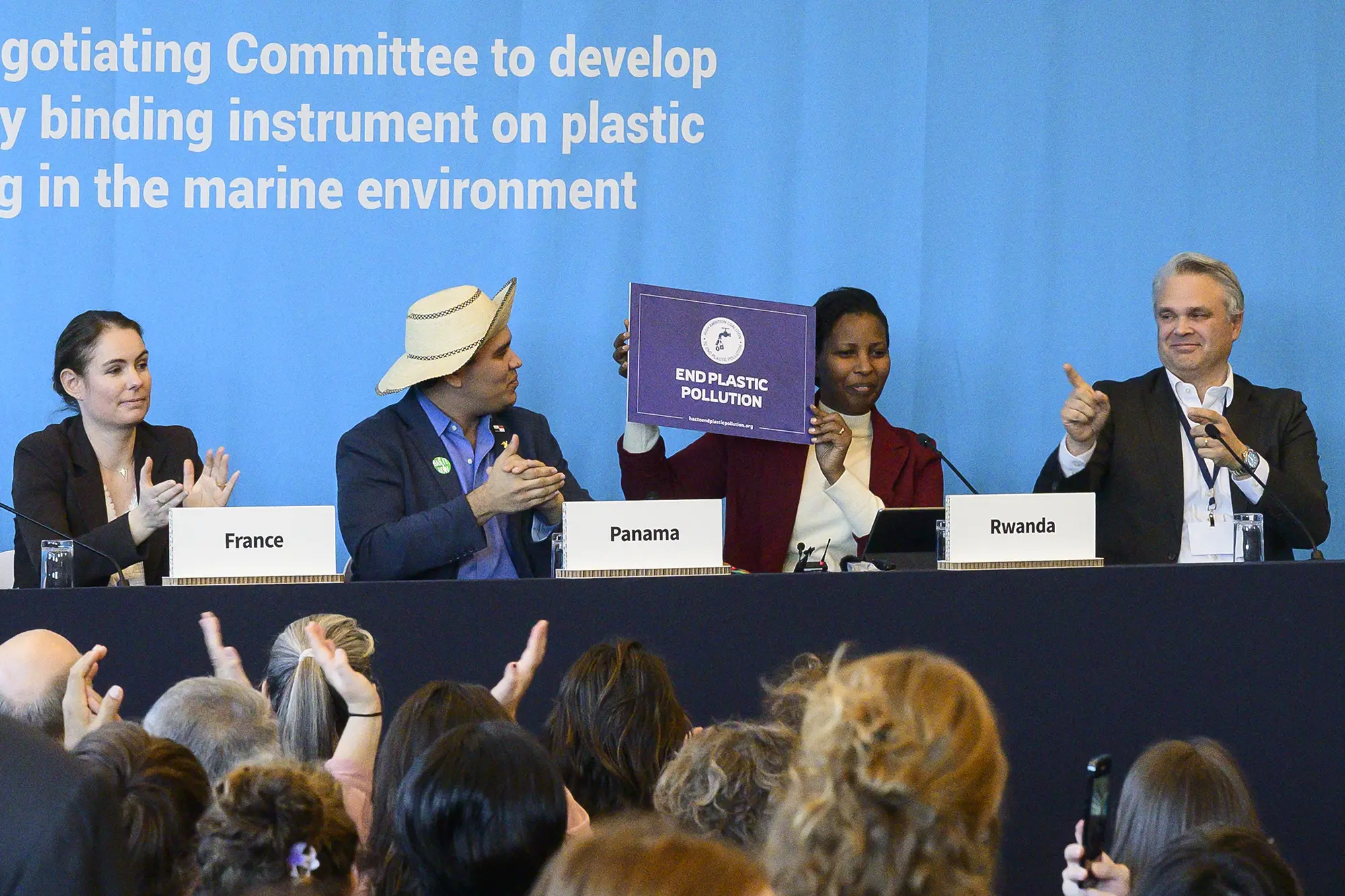 Four delegates sit at a long black covered table in front of a UN blue background. The delegates are Olga Givernet from France, Juan Carlos Monterrey from Panama, Juliet Kabera from Rwanda and Anthony Agotha representing the EU. Juliet Kabrera is holding up a purple placard that says "End Plastic Pollution", while Anthony Agotha points to it in support and the other two delegates applaud.