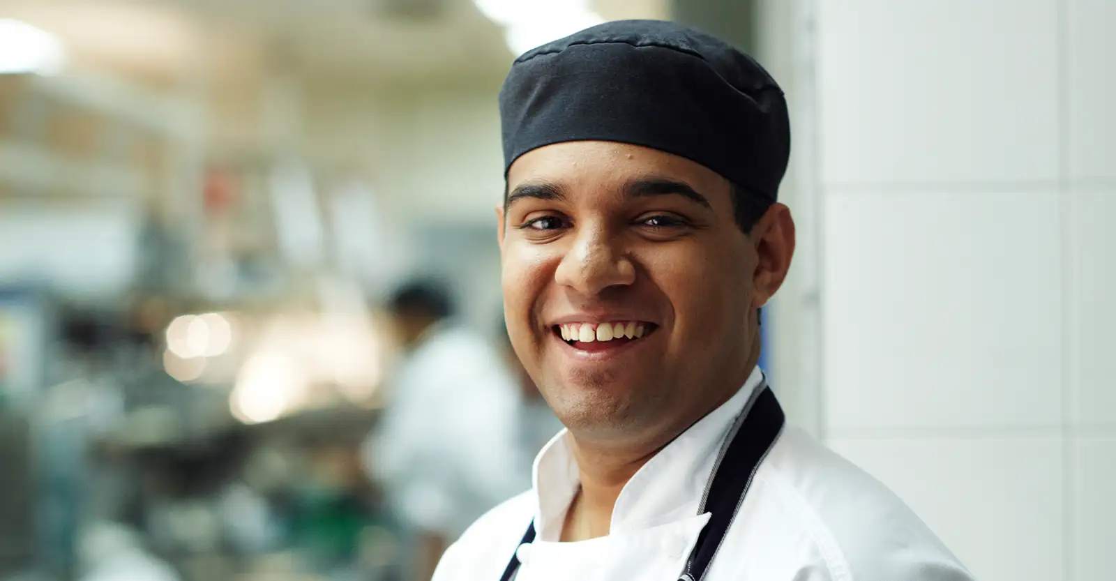 A First Nations man wearing a chef's cap and apron smiles enthusiastically into the camera. Behind him, very blurred, is a commercial kitchen.