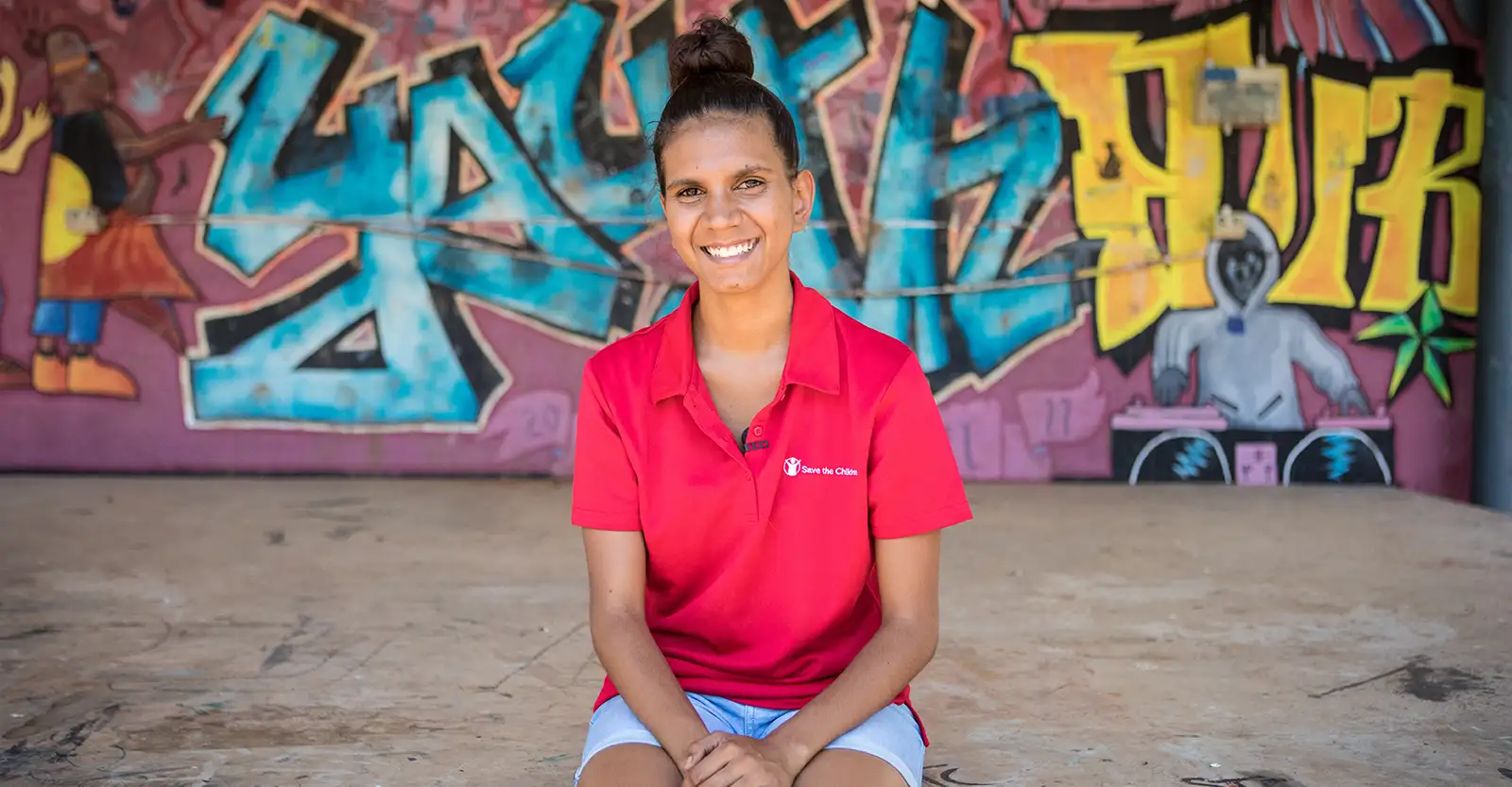 An Indigenous woman sits on a concrete stage, wearing a bright red polo shirt with the Save the Children logo on it, smiling into the camera. In the background behind her is a cool graffiti piece that says “Youth Hub”.